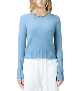 A woman wears the Shrunken Crew Cashmere Sweater by Guest in Residence in light blue, styled with white high-waisted pants and a beaded necklace.