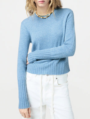 A person wears the Guest in Residence Shrunken Crew Cashmere Sweater in light blue with white pants and a multicolored beaded necklace, standing against a plain light background.