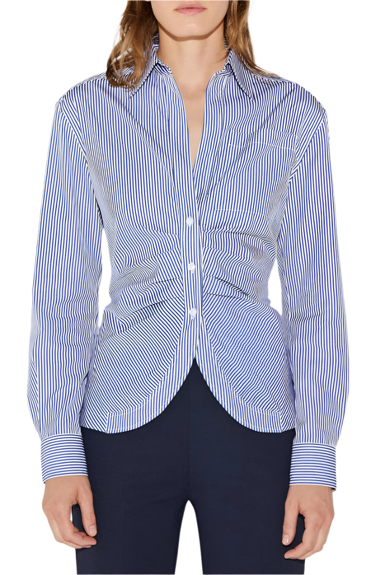 A person wearing the Sierra Top by Callas Milano, a blue and white striped poplin shirt with long sleeves, paired with dark pants. The button-front design features a gathered waist for a flattering silhouette.