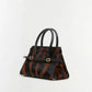 The Simkhai River Small Top Handle Bag is a structured accessory featuring black leather handles, a brown and black striped pattern, gold hardware, and a detachable crossbody strap, all showcased against a plain white background.