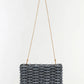 The Simkhai Morgan Clutch is a rectangular woven black and silver handbag featuring a thin, long gold shoulder strap, displayed against a plain white background.