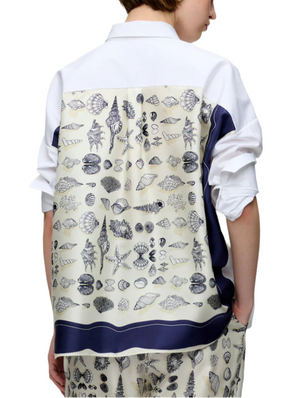 A person wears the Sirene Top by Callas Milano, a white cotton poplin shirt with a silk twill back panel showcasing seashell, starfish, and sea life prints, paired with matching pants.
