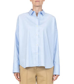 A person wearing the Sirene Top by Callas Milano, a light blue cotton poplin shirt with long sleeves, paired with beige pants and a silk twill scarf, stands against a plain white background.