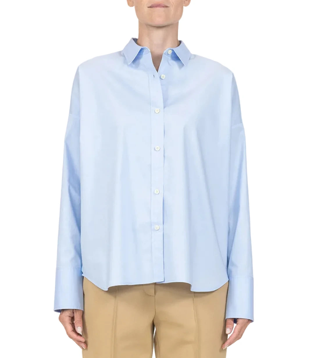A person wearing the Sirene Top by Callas Milano, a light blue cotton poplin shirt with long sleeves, paired with beige pants and a silk twill scarf, stands against a plain white background.