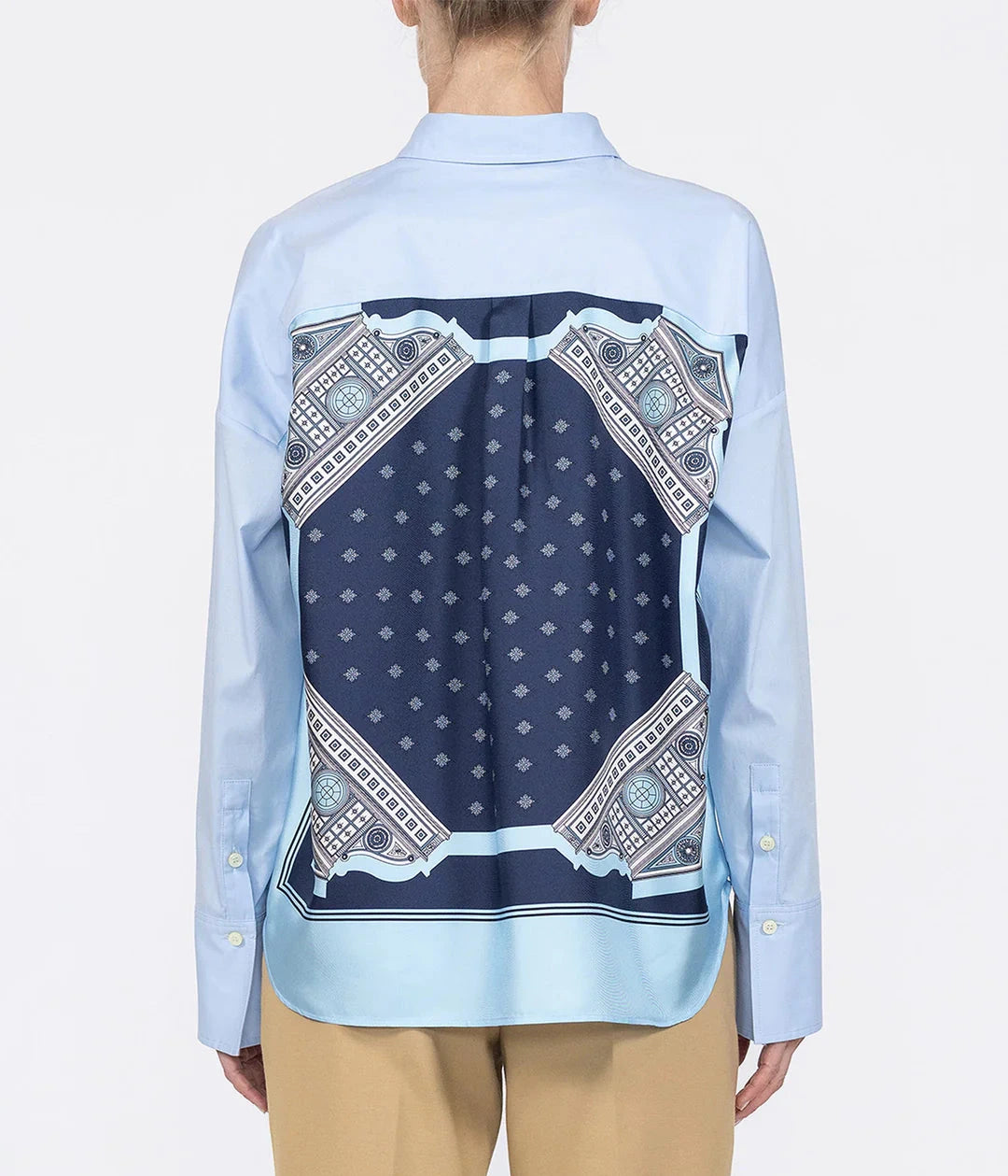 A person wears the Sirene Top by Callas Milano, a light blue cotton poplin shirt with a dark blue patterned silk twill scarf design on the back, paired with beige pants.