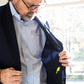 A man in a Sid Mashburn Ghost Blazer is looking at the inside pocket, which contains a green plastic dinosaur toy. He is standing near a window with bright sunlight streaming in.