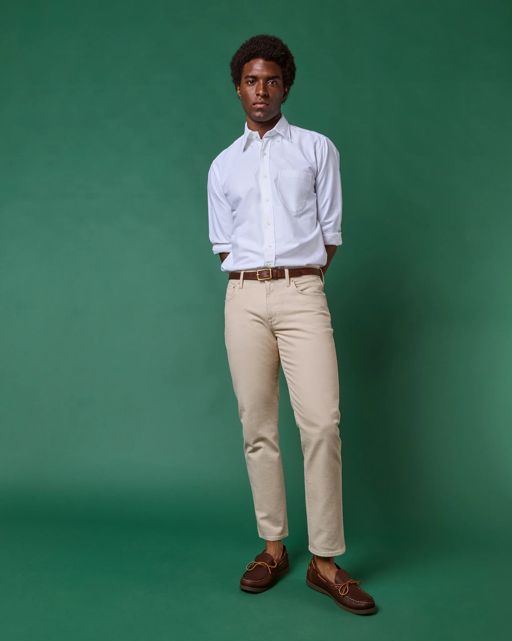 A man stands against a green background wearing a white dress shirt with rolled sleeves, Sid Mashburn Clift Straight Leg Jeans, a brown belt, and brown shoes.