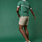 A person wearing the Sid Mashburn Rally Polo Sweater in green, along with beige shorts and white sneakers, stands against a solid green background, facing away from the camera.