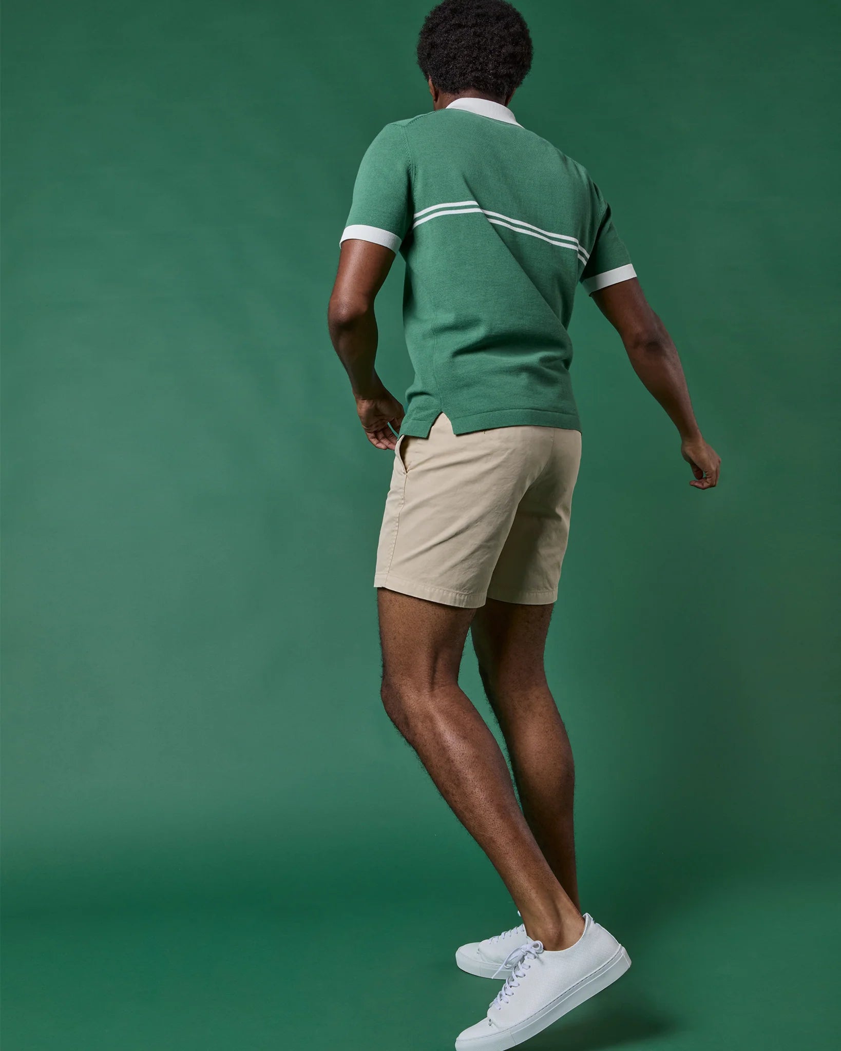 A person wearing the Sid Mashburn Rally Polo Sweater in green, along with beige shorts and white sneakers, stands against a solid green background, facing away from the camera.