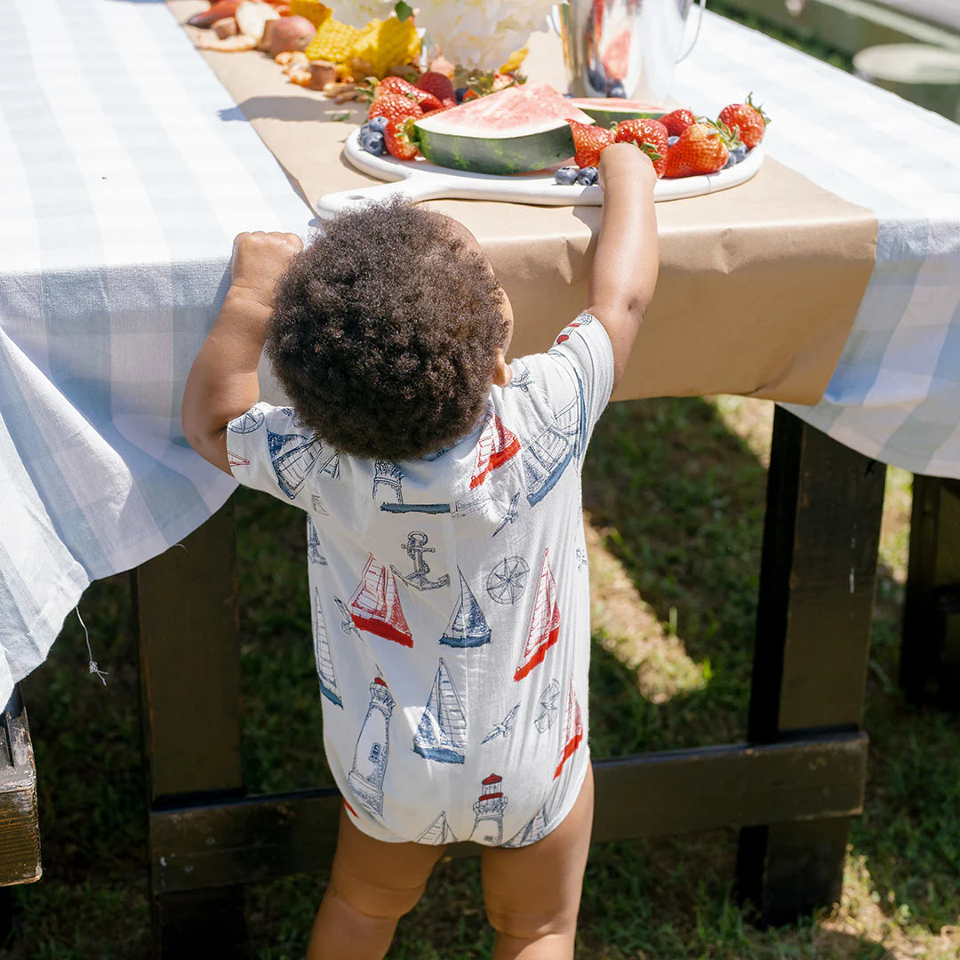 A toddler in a Pink Chicken Baby Boys' Easton Jumper with a sailboat pattern reaches for fruit on an outdoor table with a checkered tablecloth, dressed for fresh nautical adventures.