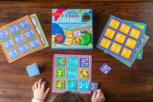 A child’s hands playing Eeboo’s Simple Math Bingo with vibrant cards and tiles displaying addition and subtraction problems on a wooden table, helping to strengthen early math skills.