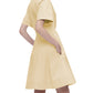 A person models the Sora Dress by Callas Milano: a knee-length, short-sleeved dress in Italian stretch poplin with a round neckline. The sleek yellow silhouette is enhanced by hands gracefully tucked in pockets while facing slightly away from the camera.
