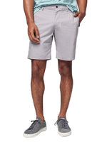 A person wearing a light blue shirt, Faherty Movement Chino Short 9" made from breathable fabric, and gray sneakers stands with hands resting on shorts. The focus is on the shorts and legs.