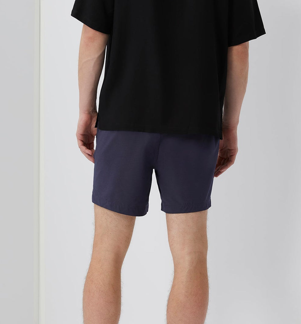 A person faces away from the camera, wearing a black T-shirt and Onia Land to Water Short 6" in navy blue with four-way stretch, against a plain background.