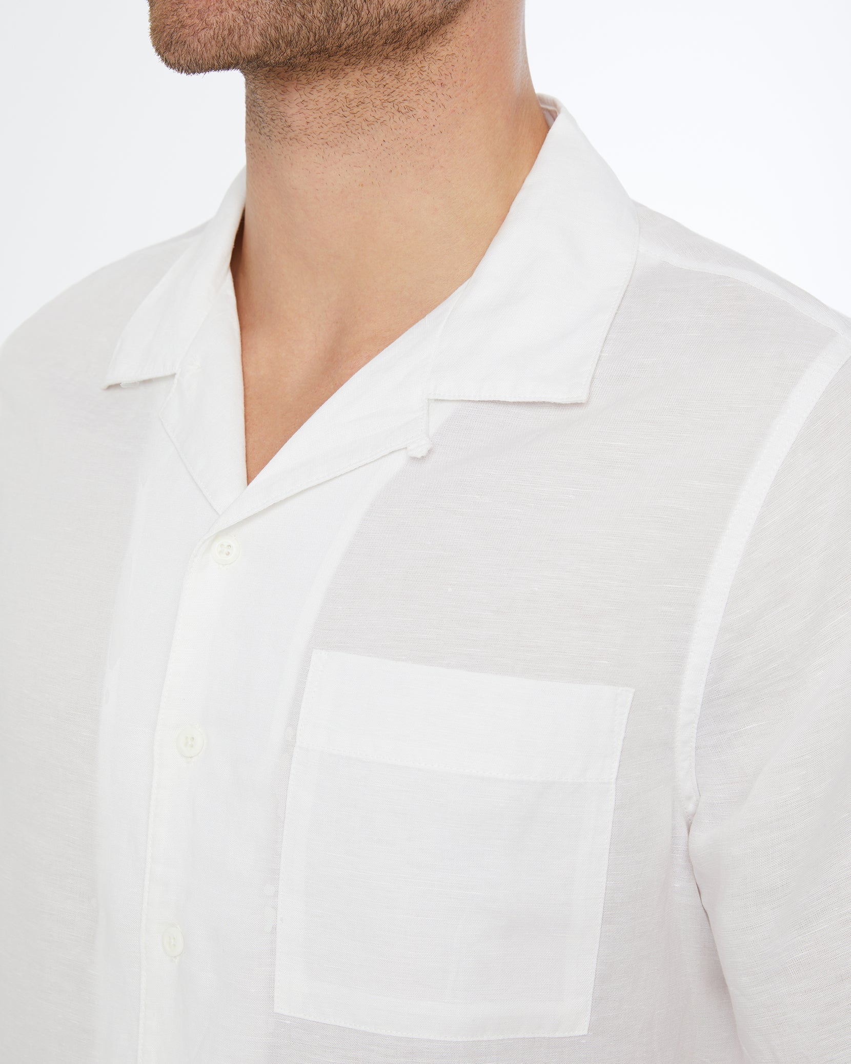 A close-up of a man wearing the Onia Air Linen Convertible Vacation Shirt, shown from shoulders to chin. The shirt features a camp collar, white linen-tencel blend, chest pocket, and open neckline.