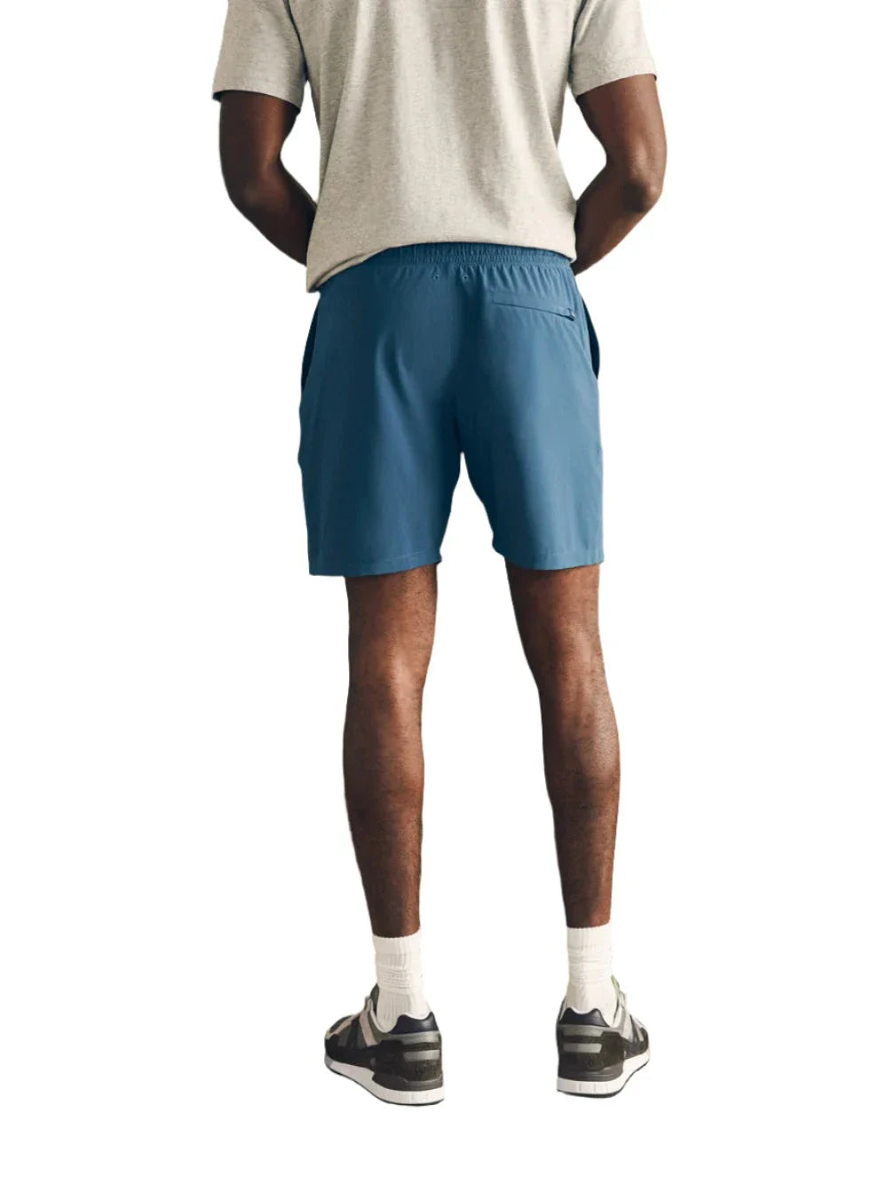 A person stands facing away, wearing a gray t-shirt and Faherty Shorelite Swim Trunks—blue athletic shorts crafted from quick-drying fabric—along with white socks and black sneakers, hands resting on their hips against a plain white background.