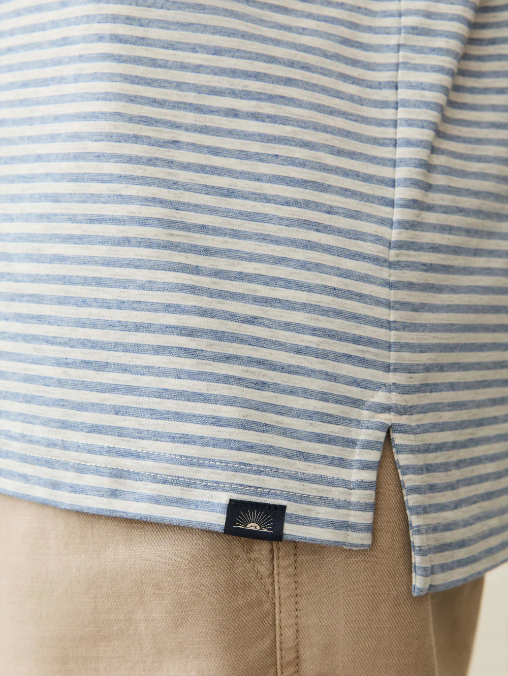 Close-up of Faherty's Short-Sleeve Movement™ Polo Shirt, featuring blue and white stripes with a small black tag above a side slit, paired with tan pants. Designed for effortless ease of movement, it combines performance qualities for comfort and style in every stride.