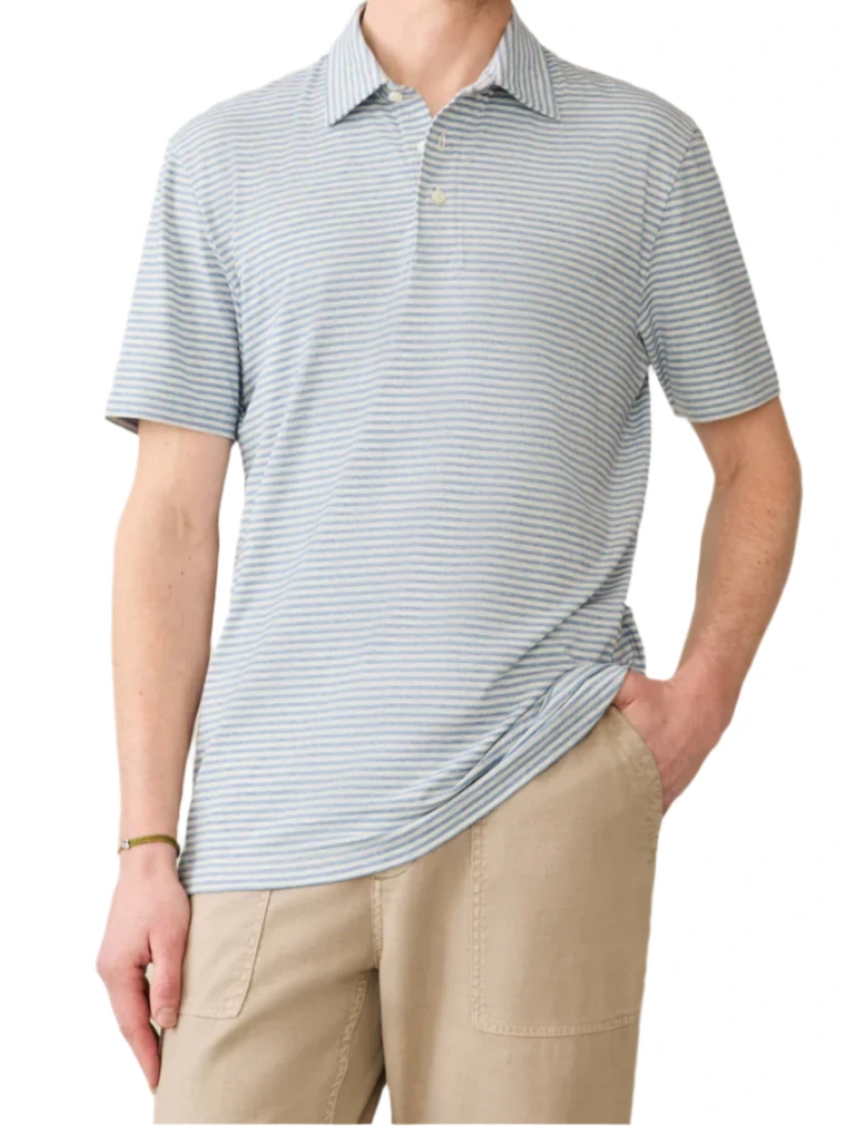 A person in a Faherty Short-Sleeve Movement™ Polo Shirt with light blue and white stripes and tan pants stands casually with one hand in their pocket, exuding effortless ease.