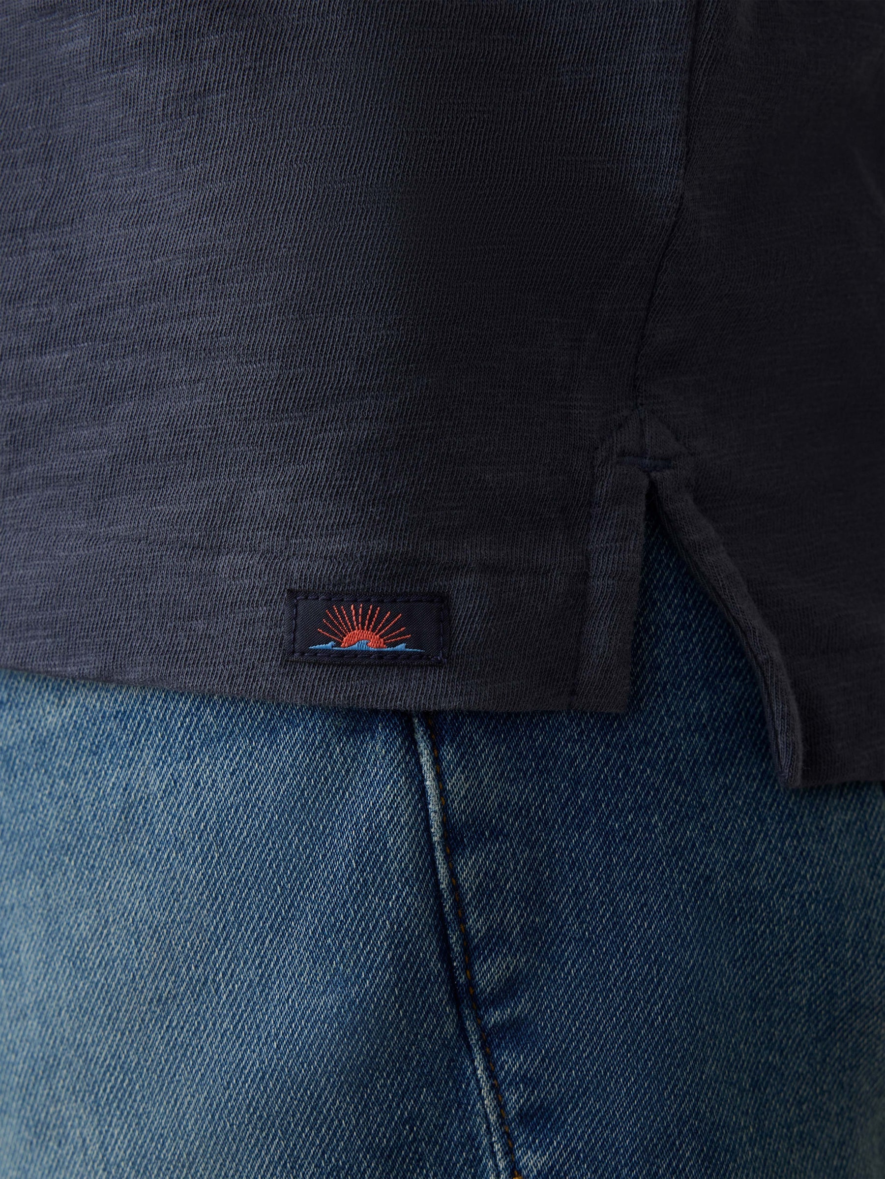 Close-up of the hem of the Faherty Sunwashed T-Shirt Polo, made from Regenerative Organic Cotton, featuring a small embroidered patch and side slit, worn over blue denim jeans.