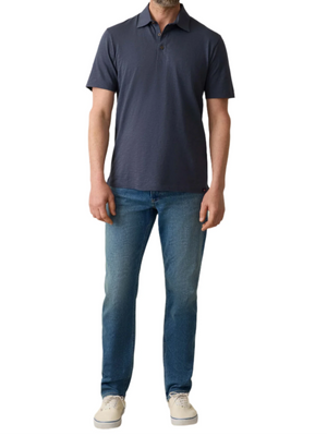 A man stands facing forward against a white background wearing a navy Sunwashed T-Shirt Polo by Faherty, blue jeans, and white sneakers.