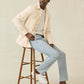 A man sits on a wooden stool against a plain background wearing the Faherty Sunwashed Canvas CPO—a relaxed fit cream shirt jacket—paired with a striped shirt, light blue pants, and white sneakers.