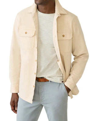 A man wears the Faherty Sunwashed Canvas CPO, a relaxed fit light beige shirt jacket, over a white and gray striped tee with light gray pants and a brown belt.