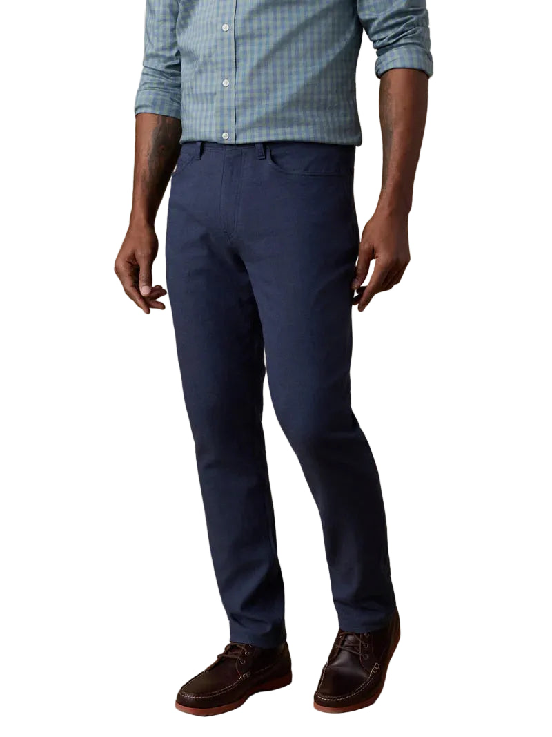 A man stands against a plain background wearing a light blue checkered shirt, brown shoes, and Faherty’s Movement Linen 5-Pocket Pant in navy blue.
