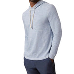 A man wears the Faherty Sunwashed Slub Hoodie, a relaxed fit, light blue cotton hoodie with white drawstrings, paired with dark blue pants and standing with his hands in his pockets.