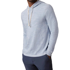 A man wears the Faherty Sunwashed Slub Hoodie, a relaxed fit, light blue cotton hoodie with white drawstrings, paired with dark blue pants and standing with his hands in his pockets.