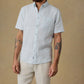 A man is wearing the Faherty SS Breeze Shirt—a light blue, lightweight linen shirt with a button-down collar and front pocket—paired with beige pants, standing against a plain tan background.