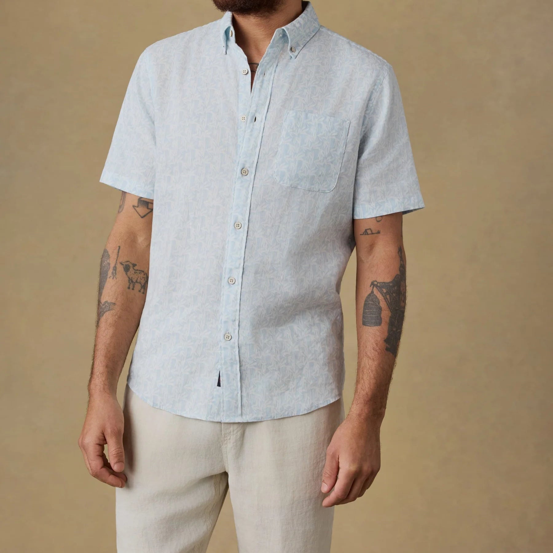 A man is wearing the Faherty SS Breeze Shirt—a light blue, lightweight linen shirt with a button-down collar and front pocket—paired with beige pants, standing against a plain tan background.