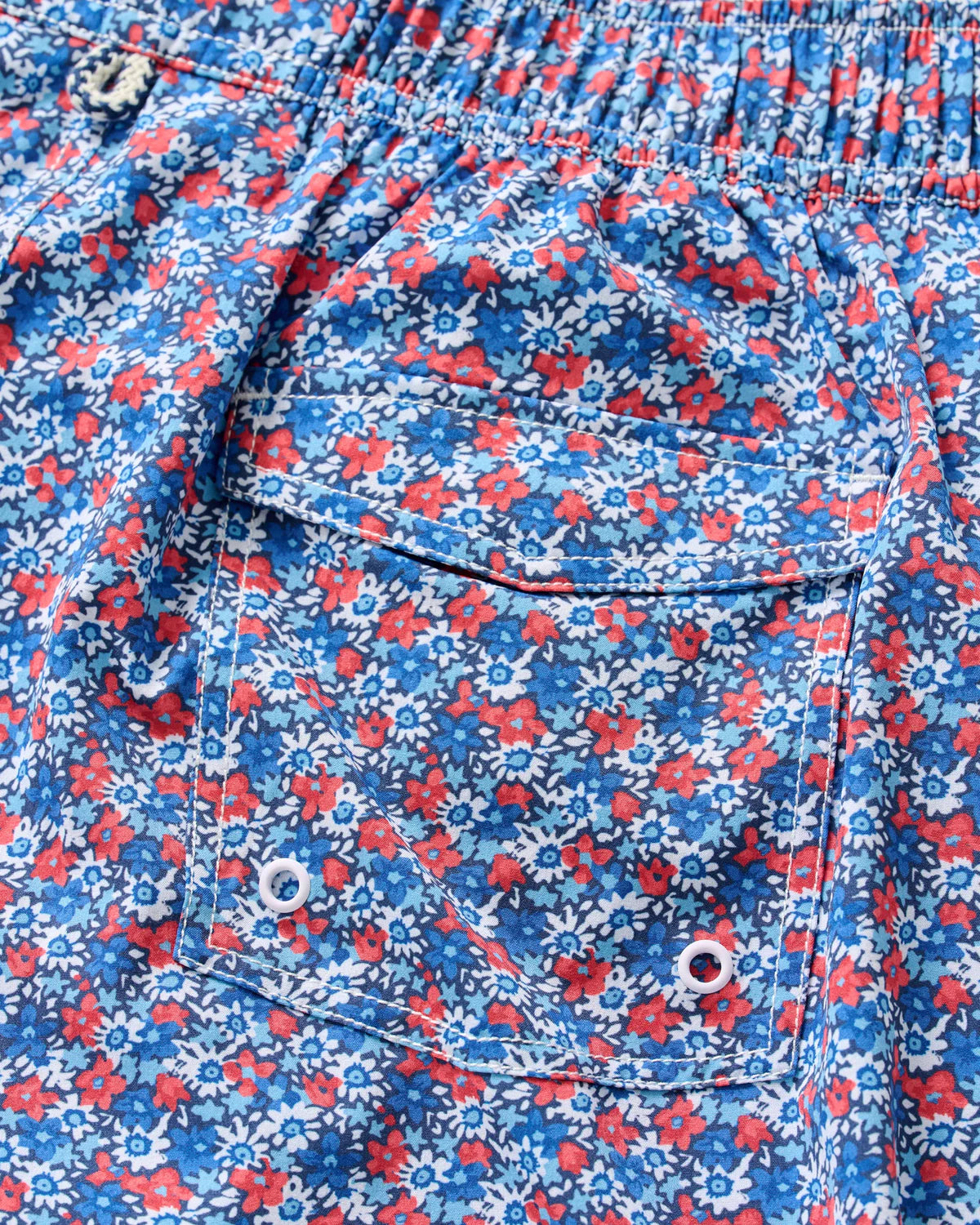 Close-up of the pocket on the Johnnie-O Freesia Swim Trunk, featuring blue fabric with a lively red, white, and blue floral print—ideal for anyone who loves fun patterns. The pocket includes metal eyelets and a buttoned flap, adding classic surf short-inspired functionality.