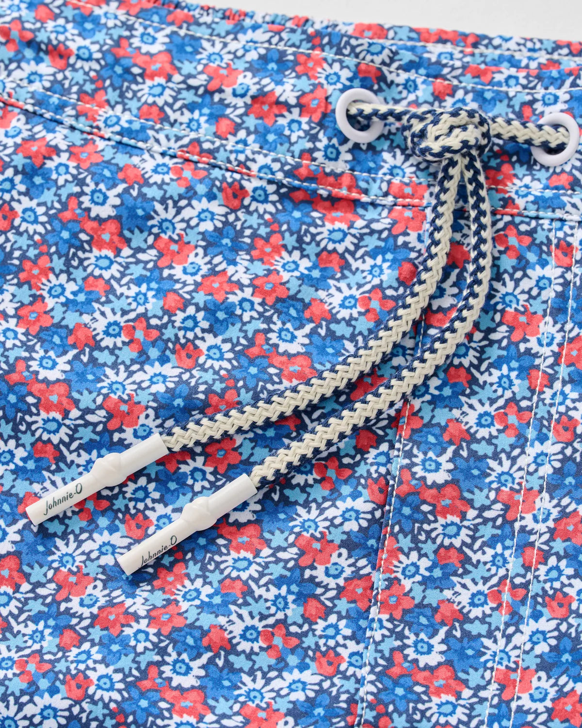 Close-up of the Johnnie-O Freesia Swim Trunk, showcasing a playful blue and red floral print, navy and white braided drawstring with metal grommets, and a 7" inseam.