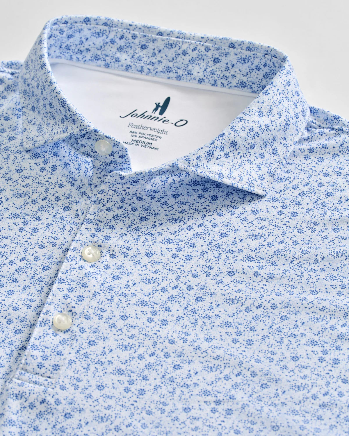 The Marietta Polo by Johnnie-O features a light blue hue with a small dark blue floral pattern, accented by white buttons and a branded label inside the collar. Crafted in a modern featherweight design, this polo delivers comfort and subtle sophistication—perfect for warm days at the office or enjoying sun protection outdoors.
