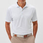 A man stands with hands in pockets, wearing the Johnnie-O Marina Polo in light blue—crafted from moisture-wicking fabric—tucked into beige pants secured with a tan belt.
