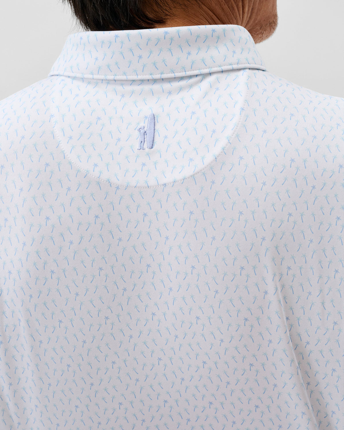 Close-up of the back of a person wearing the Johnnie-O Marina Polo, a white collared lightweight polo with a light blue abstract pattern, moisture-wicking fabric, and a small embroidered figure below the collar.
