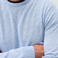 A person wearing the Johnnie-O Albus Crew—a light blue ultrasoft crewneck sweater with ribbed cuffs and collar—stands with arms crossed against a plain background, showcasing the ideal lightweight sweater for everyday layering.