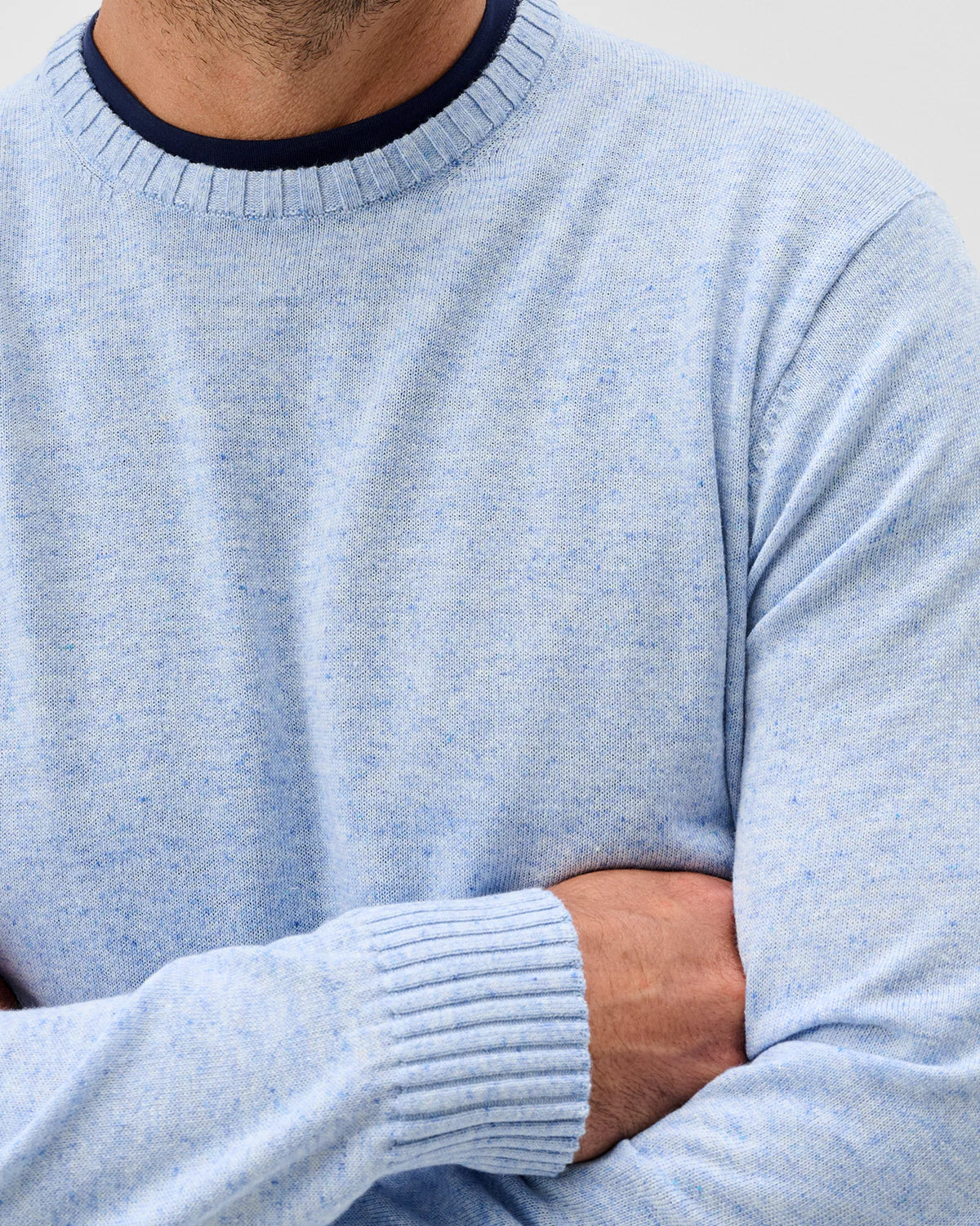 A person wearing the Johnnie-O Albus Crew—a light blue ultrasoft crewneck sweater with ribbed cuffs and collar—stands with arms crossed against a plain background, showcasing the ideal lightweight sweater for everyday layering.