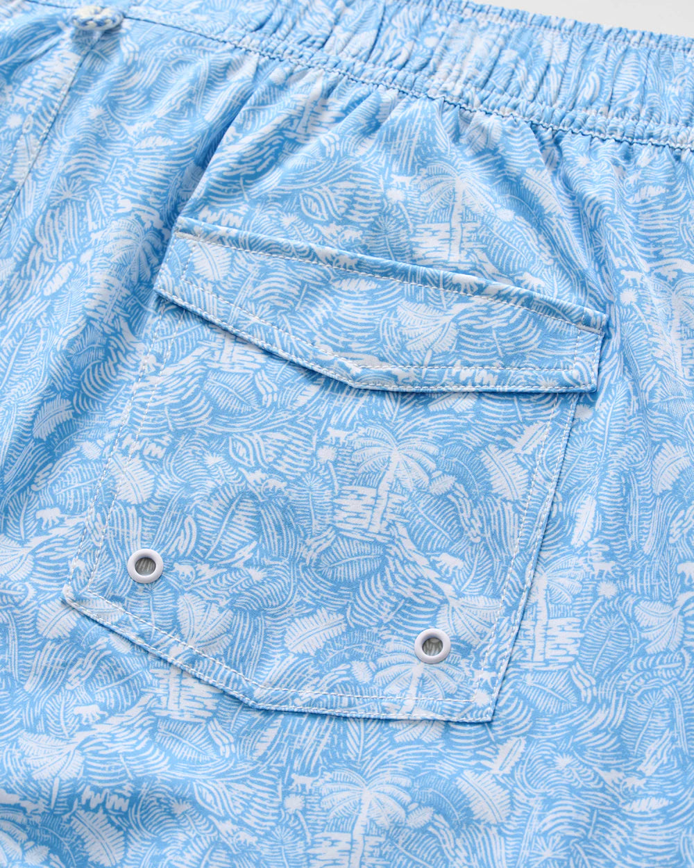 Close-up of Johnnie-O Swingin Swim Trunks, featuring a vibrant blue and white tropical pattern, an elastic waistband, and a flap pocket with metal eyelets—ideal for fans of playful prints.