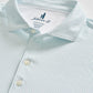 Close-up of a light blue Johnnie-O Tee Party Polo featuring a subtle geometric pattern, three white buttons, and a visible collar and tag indicating size medium. This polo offers moisture-wicking fabric and UPF 50 sun protection for all-day comfort and coverage.