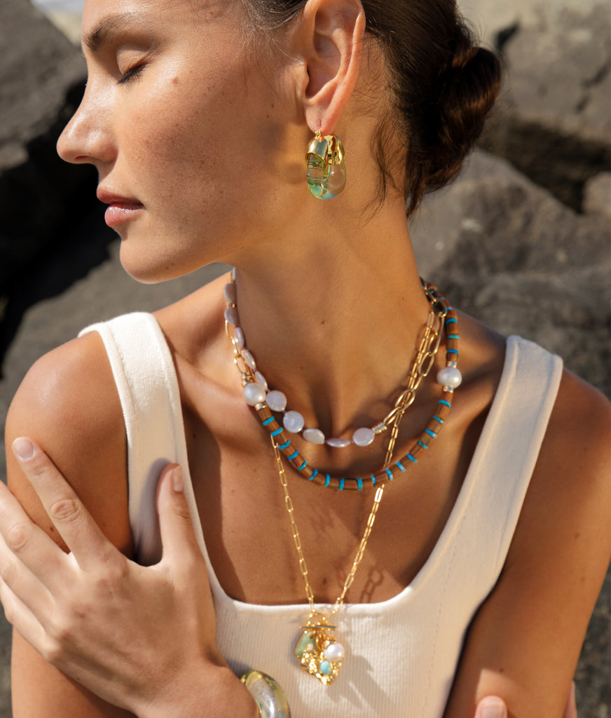 A woman, her eyes closed, stands against a rocky background, adorned with layered necklaces and wearing Lizzie Fortunato Organic Hoops.