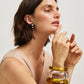 A woman with short brown hair poses in a sleeveless beige top, wearing Lizzie Fortunato’s Acacia Earrings—pearl accents with amazonite stones—as well as two chunky translucent bracelets, her hand resting near her face.