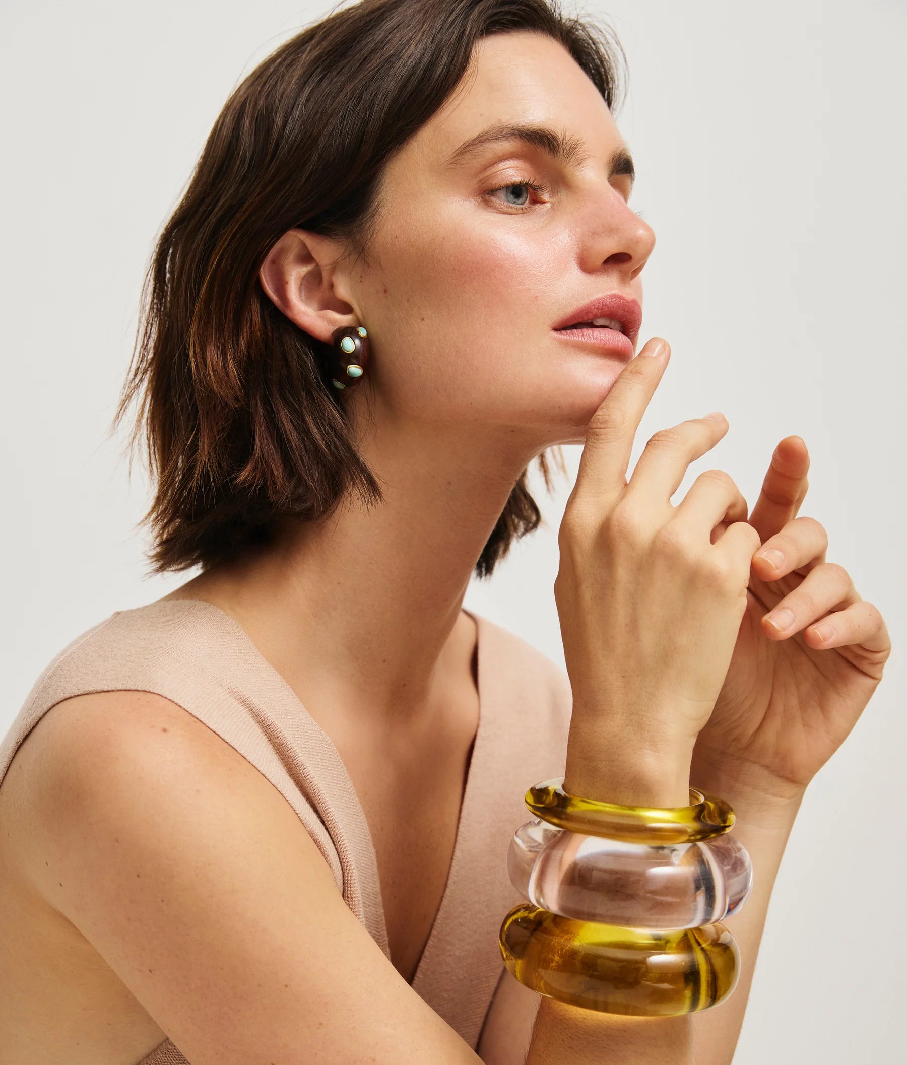 A woman with short brown hair poses in a sleeveless beige top, wearing Lizzie Fortunato’s Acacia Earrings—pearl accents with amazonite stones—as well as two chunky translucent bracelets, her hand resting near her face.