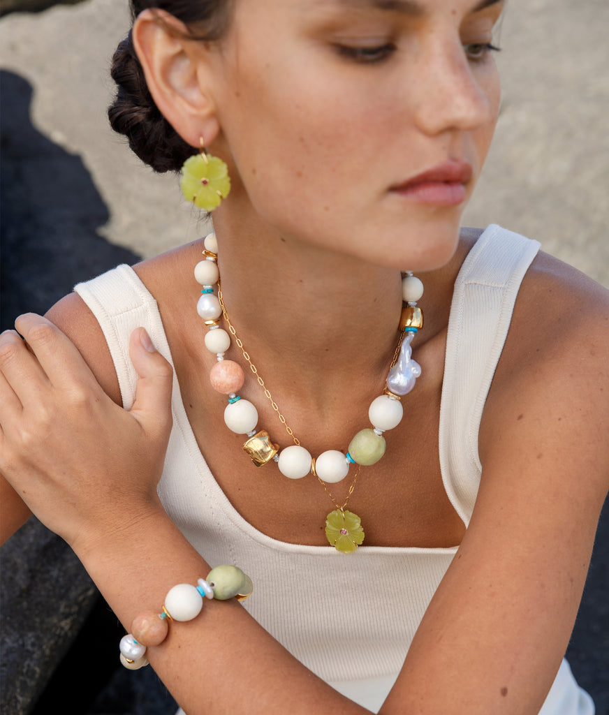 A woman wearing a white top and assorted jewelry, including the Lizzie Fortunato Andros Necklace featuring freshwater pearls and flower-shaped elements, sits gracefully on a stone surface.