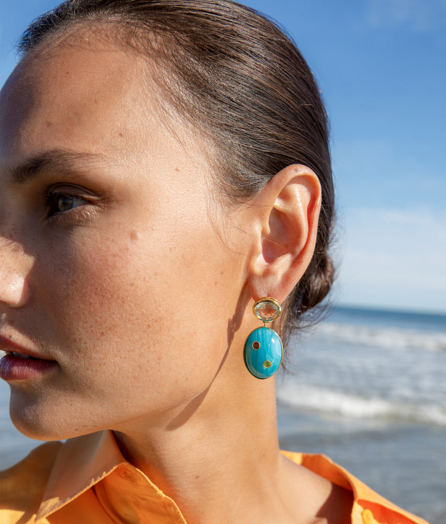 Close-up of a woman wearing Lizzie Fortunato Blue Mountain Earrings by Lizzie Fortunato, with the ocean in the background.