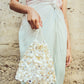 A person in a pleated pastel dress holds the Lizzie Fortunato Gala Bag, a triangular evening accessory hand-embellished with layered white and cream sequins, against a textured stone wall.