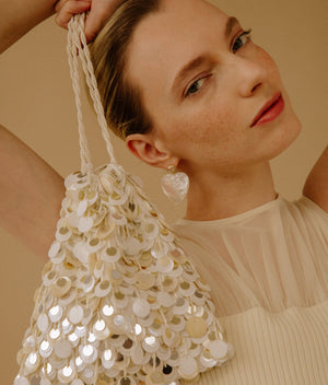 A woman holding the Lizzie Fortunato Gala Bag, a hand-embellished evening bag with large sequins, pairs it with a cream pleated top and heart-shaped earrings against a beige background.