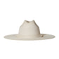 The Palmer Hat by Janessa Leone is a wide-brimmed, light beige palm straw hat featuring a pinched crown and organic cotton ties with regenerative leather aglets, photographed on a plain white background.