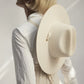 A person with short blonde hair, seen from behind in soft natural light, wears a white shirt with organic cotton ties and has the Janessa Leone Palmer Hat—a cream-colored palm straw hat—slung on their back.