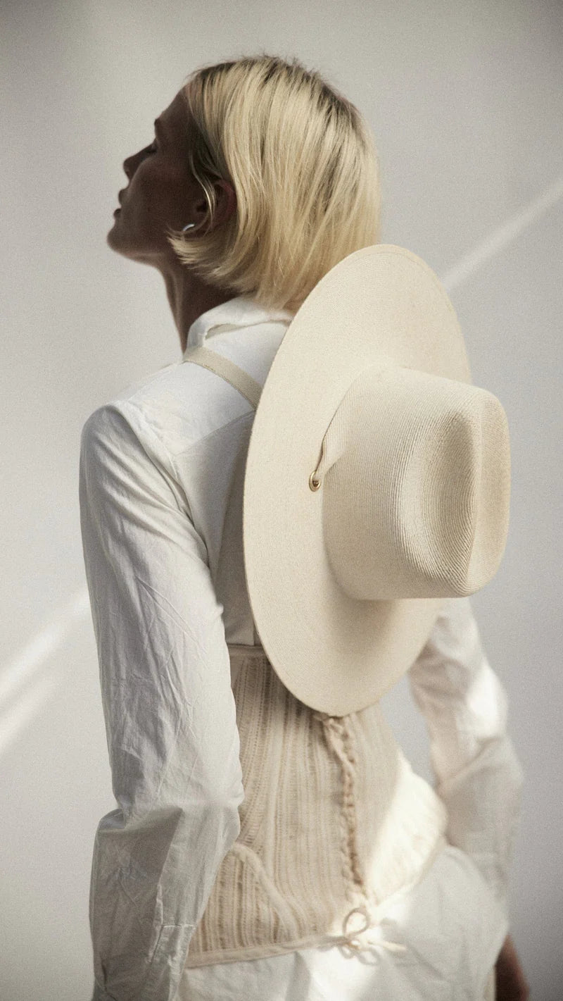 A person with short blonde hair, seen from behind in soft natural light, wears a white shirt with organic cotton ties and has the Janessa Leone Palmer Hat—a cream-colored palm straw hat—slung on their back.